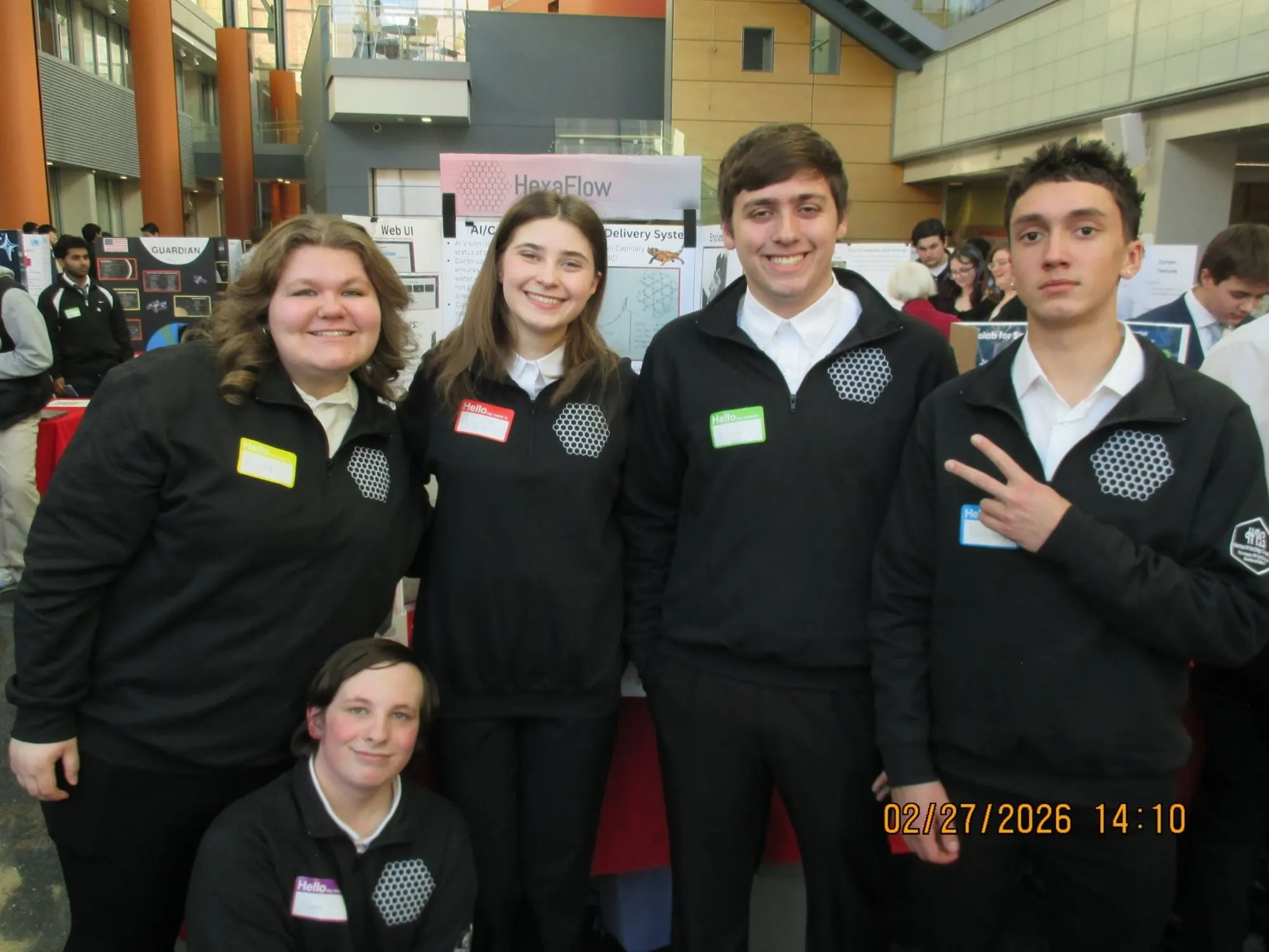 The five HexaFlow team members in matching black quarter-zips with a hexagon logo and name tags, standing in front of their HexaFlow poster at a busy science event