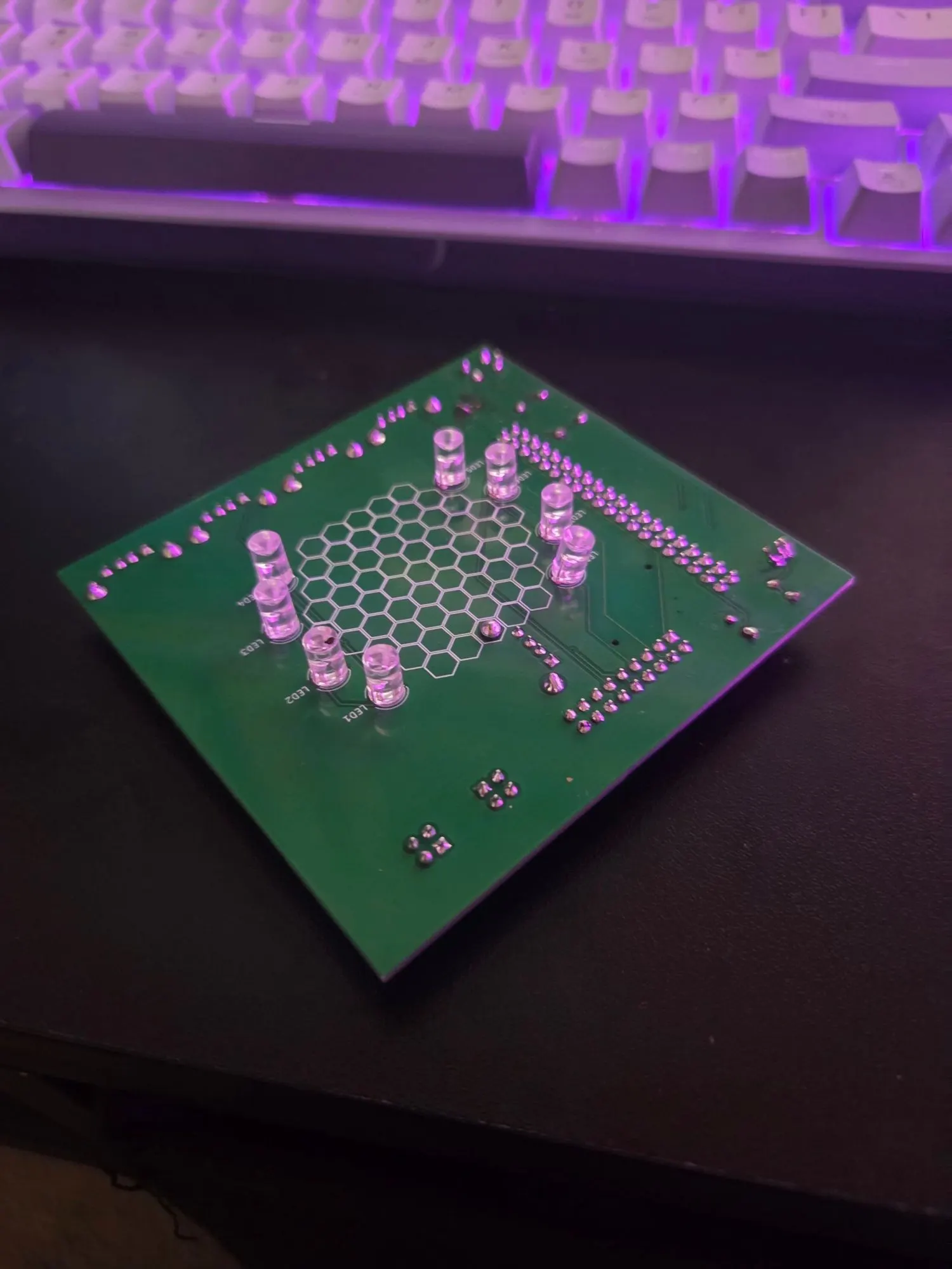 A green custom circuit board with a hexagon pattern printed on the silkscreen, ringed by clear LEDs, sitting on a desk in front of a lit keyboard