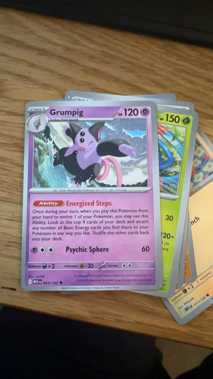 A real scan: a Grumpig card with a light silver border lying on a light wooden desk, more cards fanned out underneath it.